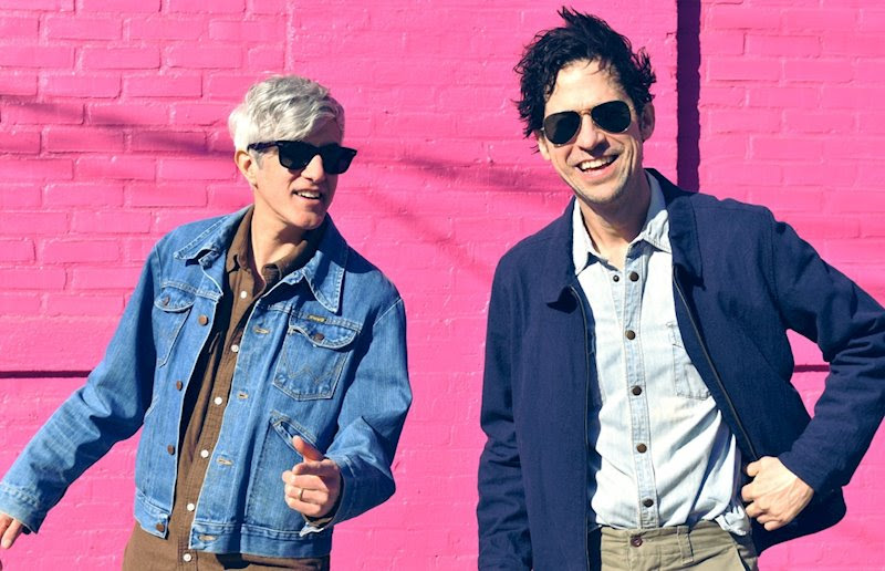 Band photo of We Are Scientists standing against a pink wall